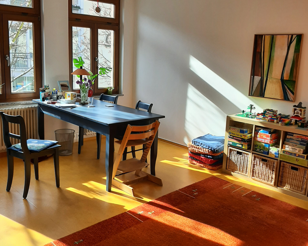 Heller Therapieraum der Logopädie-Praxis mit einem blauen Holztisch, Stühlen (inklusive Kinder-Therapiestuhl) und einem Regal voller Therapiematerialien.