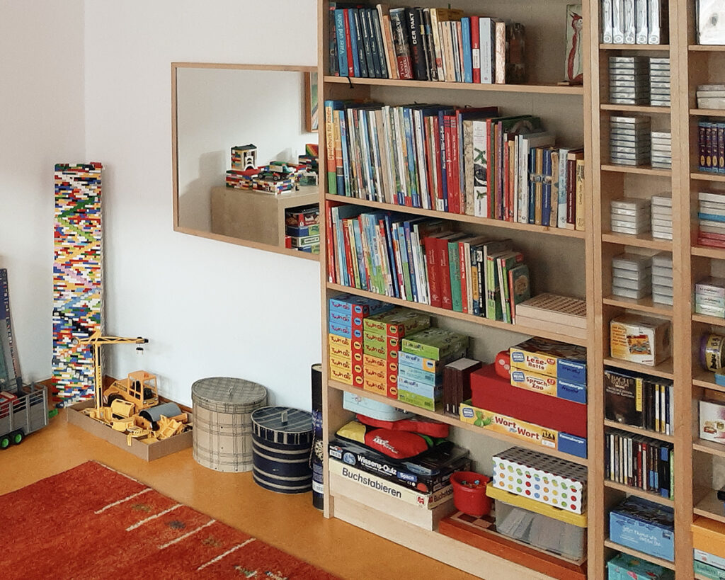 Ein hohes Holzregal gefüllt mit Büchern, Medien und Therapiematerialien, daneben eine Bauecke mit Lego-Bausteinen und einem großen Spiegel.