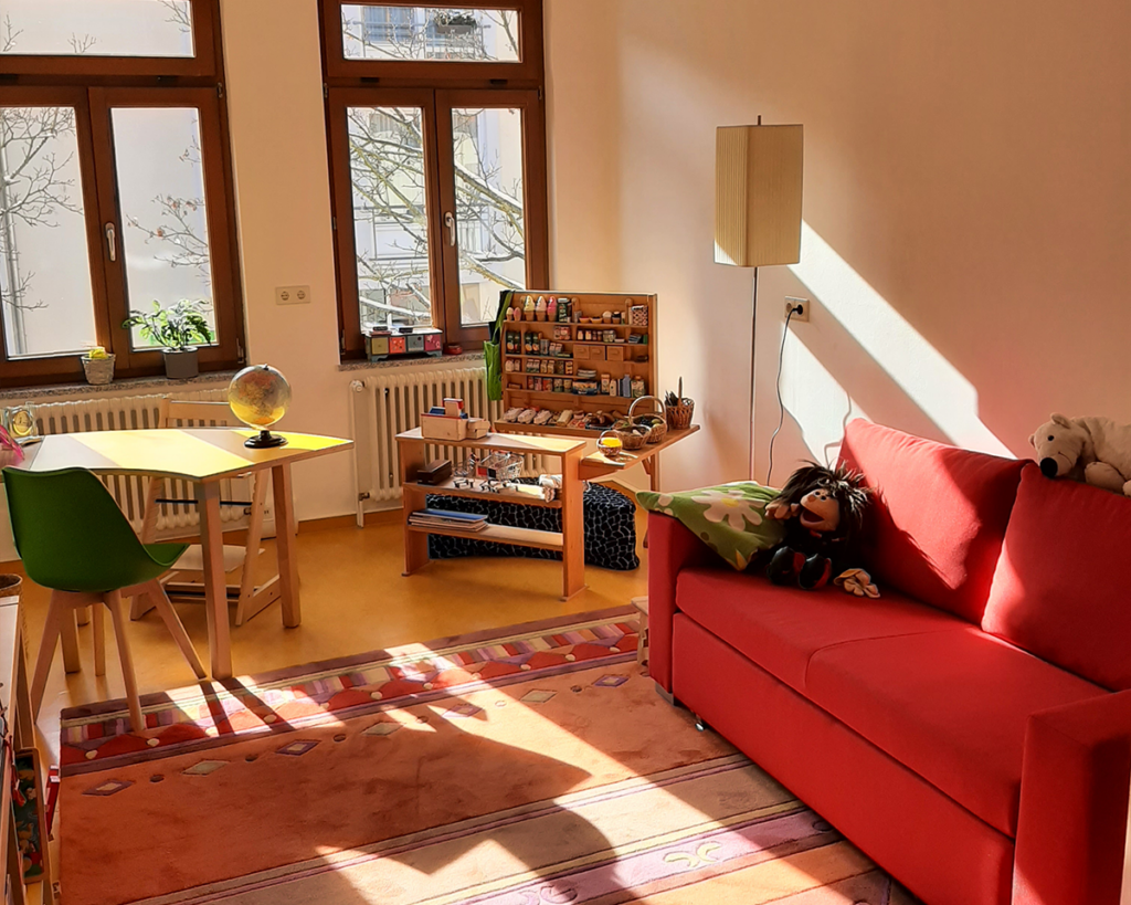 Lichtdurchfluteter Therapieraum mit rotem Sofa, Kinderspieltisch und einem gut ausgestatteten Kaufladen aus Holz zur Sprachförderung.