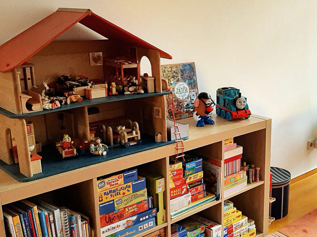 Ein Regal in der Logopädie-Praxis mit einem großen hölzernen Puppenhaus und vielen Gesellschaftsspielen zur logopädischen Förderung.