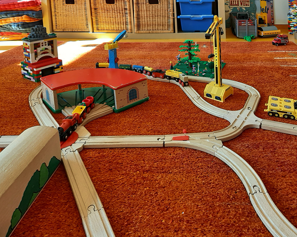 Eine große Spielzeugeisenbahn aus Holz mit Schienen und einem Lokschuppen auf dem roten Teppich des Therapiezimmers zur spielerischen Therapie.